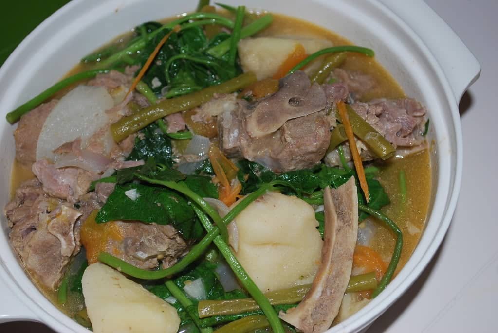 "A close-up view of Sinigang Na Ulo ng Tanigue, a traditional Filipino sour soup. The dish features chunks of tanigue (mackerel) head, fresh vegetables like water spinach, radish, and string beans, all simmered in a light, tangy broth served in a white bowl.