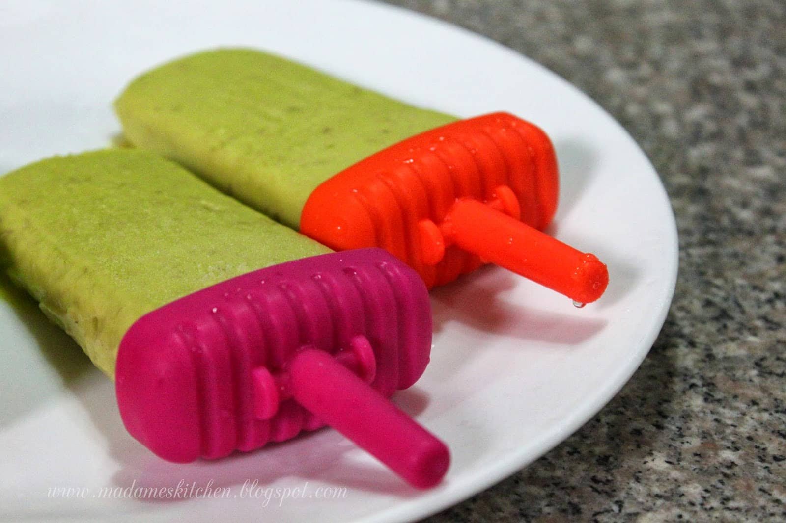Two avocado graham popsicles with colorful handles on a white plate.