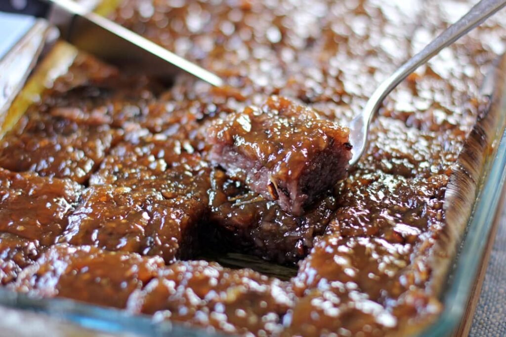 A tray of Biko, a traditional Filipino sweet sticky rice dessert, topped with a rich caramelized coconut syrup. A piece is being served, showcasing the glistening and gooey texture of the rice, with its deep brown color from the sweet topping.