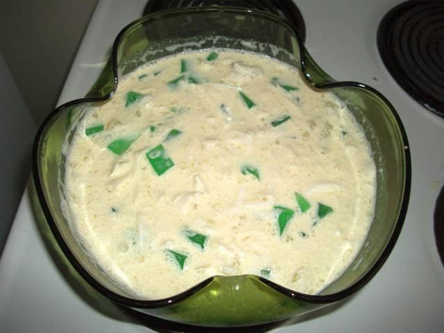 A bowl of Buko Salad, a popular Filipino dessert made from young coconut strips mixed with condensed milk, cream, and a variety of other ingredients such as jelly cubes and fruit. The creamy texture and green gelatin pieces are visible, floating in the mixture, giving the dish a festive and indulgent appearance.