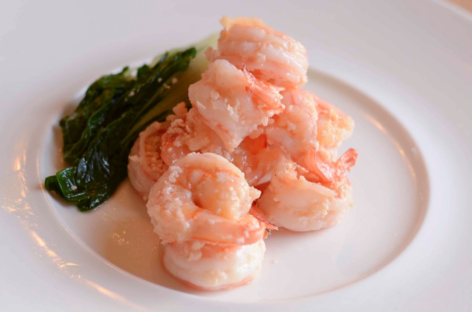 A delicious serving of butter shrimp cooked to a light pink hue, tender and glistening with a buttery glaze. The shrimp are neatly piled on a white plate, paired with a side of steamed leafy greens. The dish is simple yet elegant, showcasing the shrimp's plumpness and vibrant color