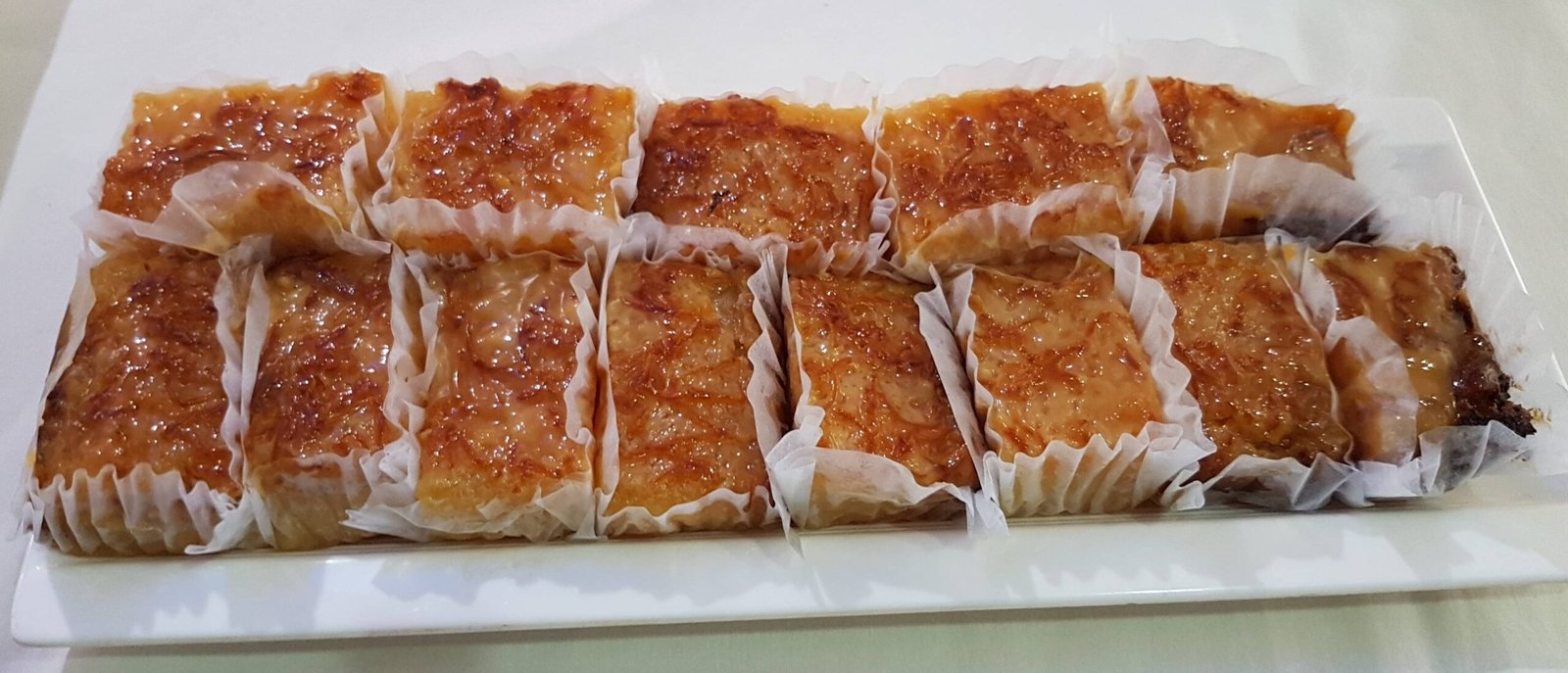 A tray of freshly baked cassava cake with macapuno, sliced into individual portions and wrapped in paper, showcasing the golden caramelized top and soft, chewy texture of this Filipino dessert.