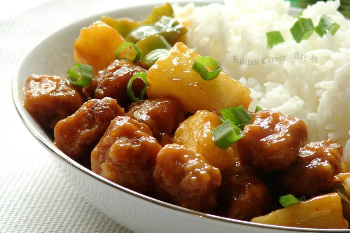 The image shows Chicken Adobo with Pineapple, a Filipino dish featuring tender chicken pieces cooked in a savory adobo sauce and garnished with chunks of pineapple. The dish is served alongside a portion of white rice and sprinkled with chopped green onions, adding a burst of color and freshness.