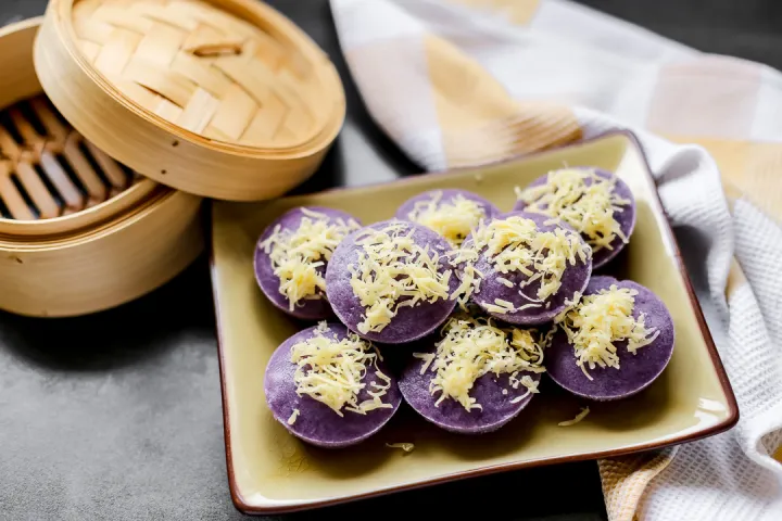 A plate of vibrant purple choco puto topped with shredded cheese, a delicious steamed Filipino rice cake dessert, served beside a bamboo steamer, perfect for trying out choco puto cheese recipes.