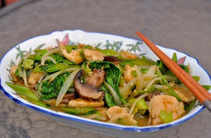 The image shows a vibrant plate of Chopsuey, filled with mixed vegetables like mushrooms, green beans, bok choy, and bean sprouts, along with pieces of chicken, served with chopsticks.