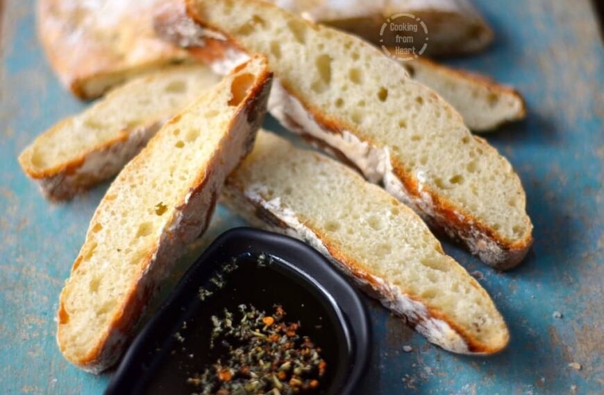 Several slices of freshly baked Broas, also known as ladyfinger biscuits, arranged on a rustic surface. The Broas are light and airy, with a crisp texture, perfect for dipping into a small dish of olive oil infused with herbs and spices.