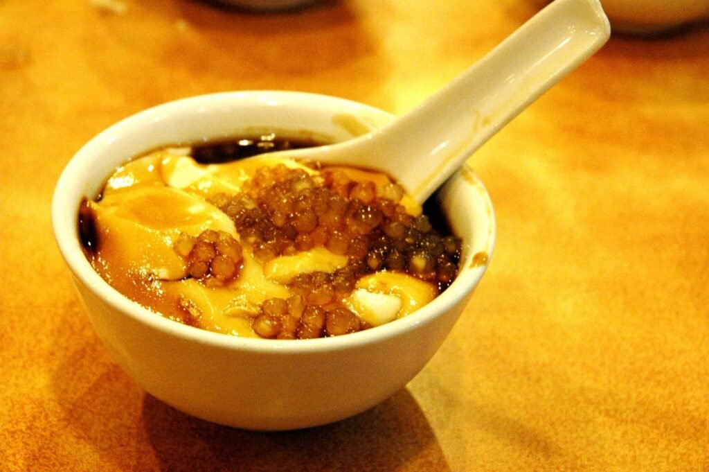 A warm bowl of Taho, a popular Filipino snack, consisting of silken tofu topped with sweet syrup and tapioca pearls. The dish is served with a small spoon, ready to be enjoyed for its smooth texture and sweet, comforting flavors.