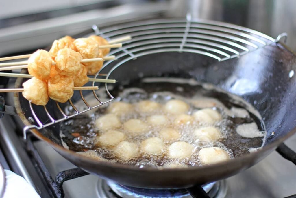 Golden brown fish balls frying in a wok, with skewers of freshly cooked fish balls resting on the side, ready to be served with Manong’s special sweet and spicy tamis-anghang sauce.