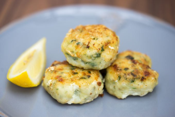 Three golden-brown fish cakes stacked on a gray plate, garnished with herbs, and served with a lemon wedge on the side, offering a light and appetizing presentation.