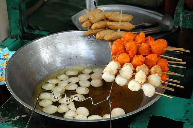 The image shows a typical Filipino street food scene with skewers of fish balls and kikiam. These are deep-fried in a large pan of oil and commonly served with various sauces, making them a popular and affordable snack in the Philippines.