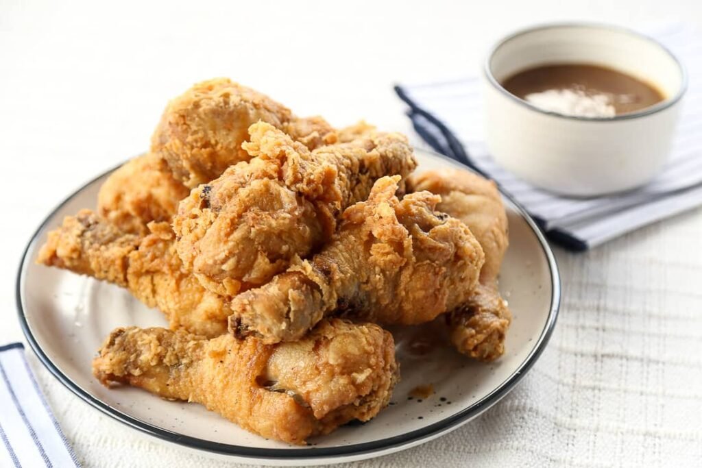 Crispy golden Fried Chicken pieces served on a white plate, reminiscent of the famous Jollibee Chicken Joy, paired with a small bowl of brown gravy on the side. The chicken is perfectly fried with a crunchy coating, displayed on a simple white tablecloth with a striped napkin, evoking the comfort and indulgence of a well-loved Filipino fast-food favorite.