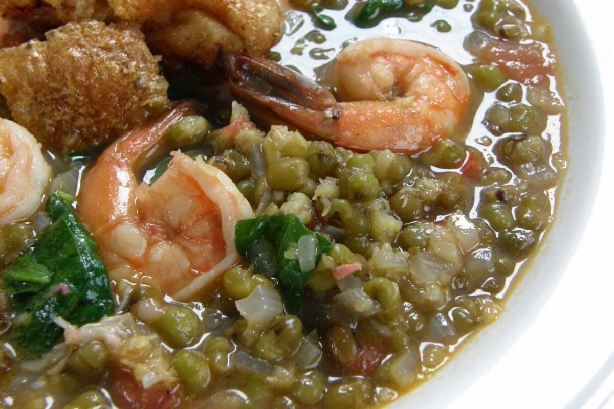 A close-up view of Ginataang Kangkong, a savory Filipino dish made with fresh shrimp, crispy pork, and mung beans in a coconut milk-based broth. The dish features water spinach (kangkong) adding a leafy texture to the mix.