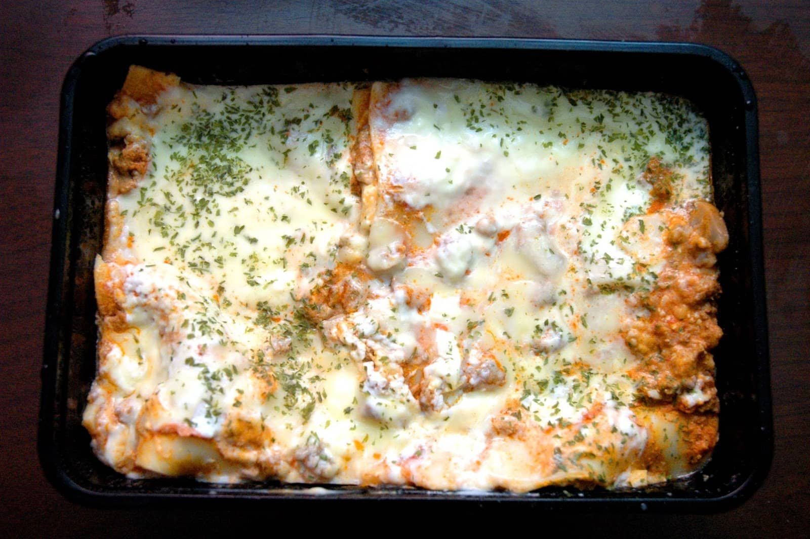 A freshly baked Greenwich-Style Lasagna Supreme topped with melted cheese and sprinkled with herbs, served in a black tray.