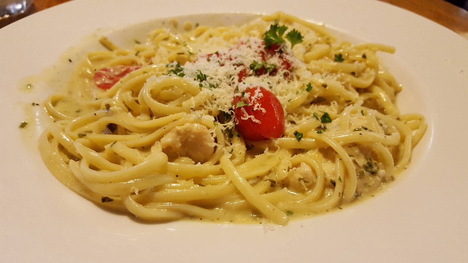 The image shows a plate of homemade carbonara, featuring creamy linguine pasta with cherry tomatoes, grated cheese, and garnished with fresh parsley. The sauce looks rich and cheesy, making for a delicious and comforting meal.