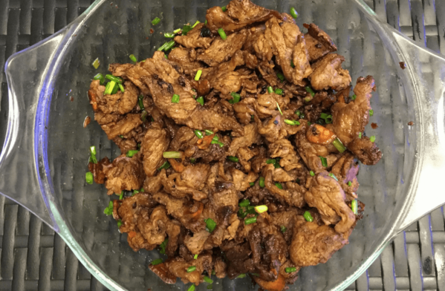 A clear glass dish filled with Homemade Pork Tapa, a Filipino dish made of marinated and fried pork slices. The pork is caramelized and cooked to a tender, slightly crispy finish. The dish is garnished with chopped green onions, adding a fresh contrast to the rich, savory-sweet marinated pork. The tapa is often served with rice, making it a popular breakfast or meal option.