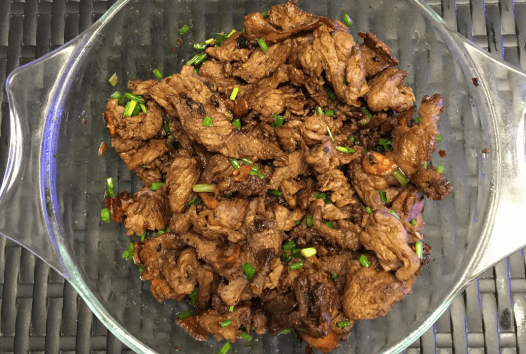 A clear glass dish filled with Homemade Pork Tapa, a Filipino dish made of marinated and fried pork slices. The pork is caramelized and cooked to a tender, slightly crispy finish. The dish is garnished with chopped green onions, adding a fresh contrast to the rich, savory-sweet marinated pork. The tapa is often served with rice, making it a popular breakfast or meal option.