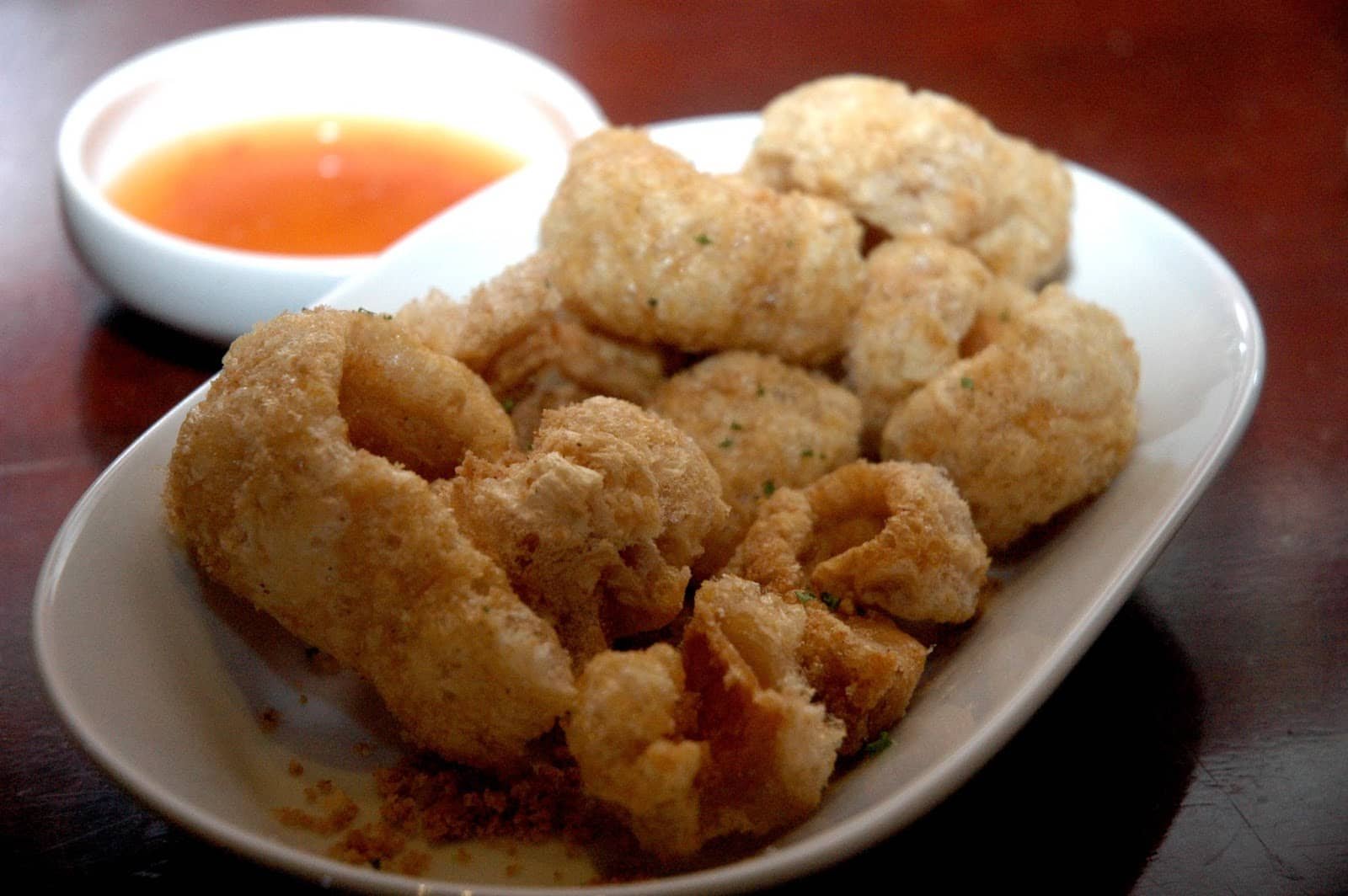 A plate of crispy fried chicharon pieces, golden and puffed, served alongside a small dish of vibrant orange Sukang Cebu vinegar dipping sauce. The crispy snack is arranged in an oval-shaped white plate, with the chicharon showing a light and crunchy texture. The vinegar dip in the background adds a bright contrast, complementing the rich and savory taste of the chicharon.