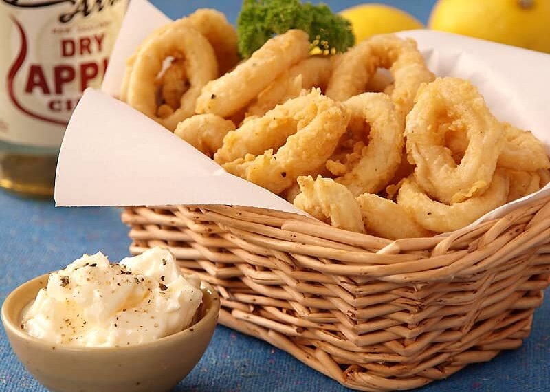 A wicker basket filled with golden-brown, deep-fried calamari rings, accompanied by a small dish of creamy dipping sauce garnished with cracked black pepper. A bottle of dry apple cider and fresh lemons are visible in the background, adding a refreshing touch to the presentation.