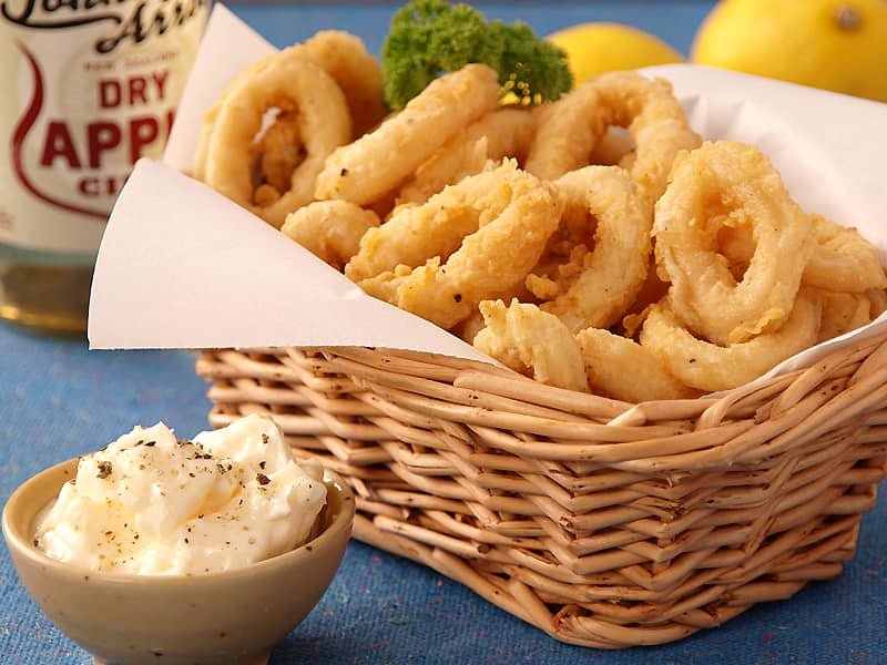 A wicker basket filled with golden-brown, deep-fried calamari rings, accompanied by a small dish of creamy dipping sauce garnished with cracked black pepper. A bottle of dry apple cider and fresh lemons are visible in the background, adding a refreshing touch to the presentation.