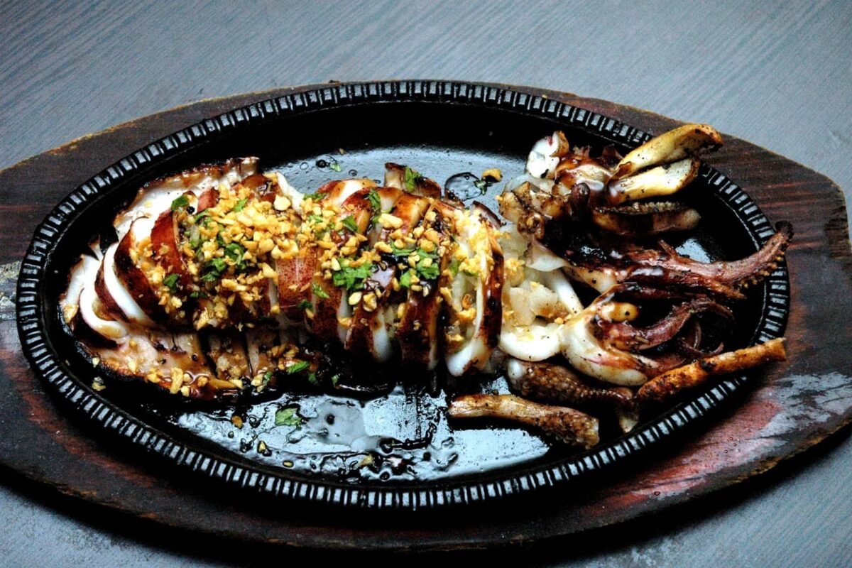 A beautifully grilled squid served on a hot sizzling plate, garnished with a generous amount of toasted garlic, chopped parsley, and a drizzle of savory sauce. The squid is tender and slightly charred, with the tentacles positioned alongside the body, enhancing the presentation. The dish is set on a rustic wooden platter, making it both visually appealing and appetizing.