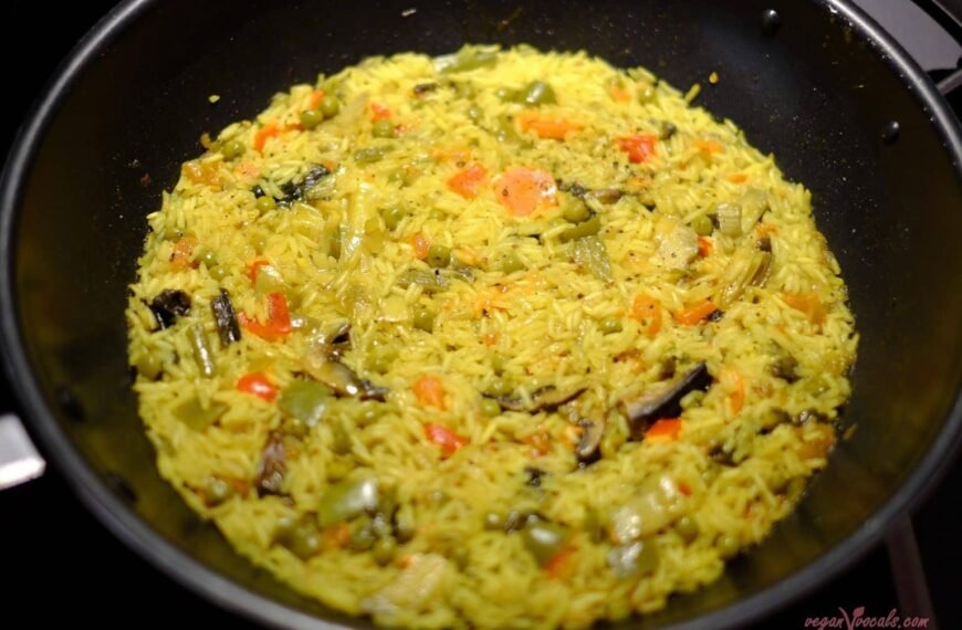 An overhead view of a colorful paella cooking in a pan. The dish features vibrant yellow rice, mixed with vegetables such as carrots, green beans, and peas, giving the paella a visually appealing combination of colors. The rice is evenly spread across the pan, cooked to perfection with a slightly glossy texture.