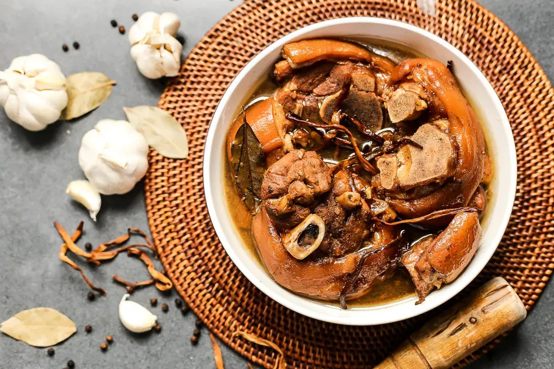 A bowl of Paksiw na Pata, a Filipino dish featuring pork hocks stewed in a flavorful mixture of vinegar, soy sauce, and spices, garnished with bay leaves, peppercorns, and garlic cloves, served on a woven mat.