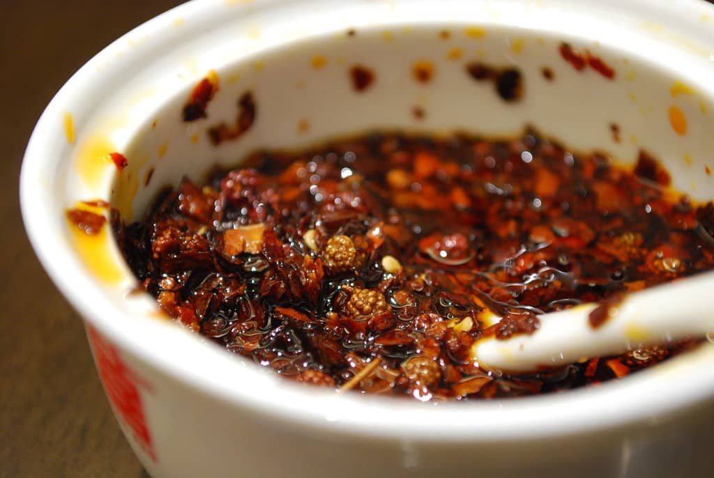 A close-up shot of a bowl filled with homemade chili garlic oil, featuring deep red chili flakes and bits of crispy garlic submerged in oil. The vibrant red hue of the oil reflects the spicy and flavorful nature of the sauce.