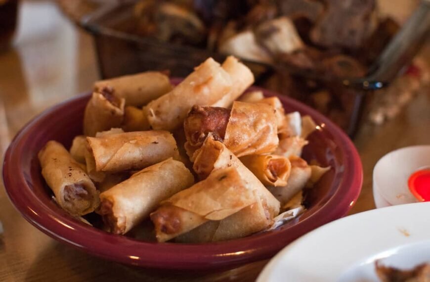 A plate filled with freshly cooked Lumpiang Shanghai, crispy and golden brown, served in a deep red bowl. Each spring roll is tightly wrapped and fried to perfection, showcasing their crunchy texture. The background shows a dining table with other dishes, enhancing the warm and inviting ambiance of a Filipino meal gathering.