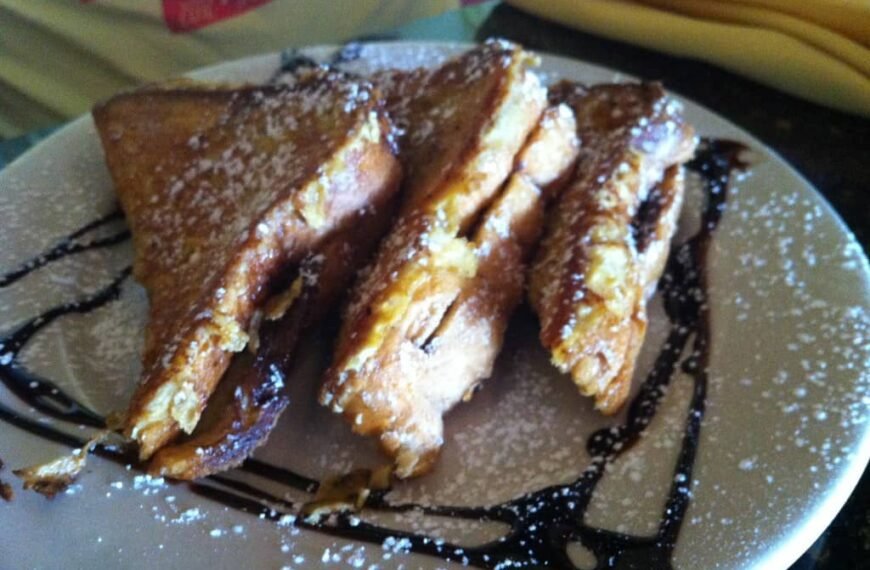 Three slices of Nutella French Toast Rolls, dusted with powdered sugar and drizzled with chocolate sauce, arranged on a white plate. The golden-brown toast is crispy on the outside, with a rich Nutella filling oozing out, making for a decadent and delicious breakfast treat.