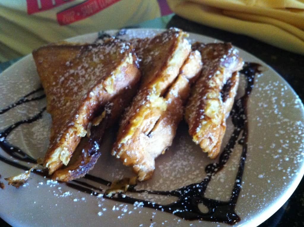 Three slices of Nutella French Toast Rolls, dusted with powdered sugar and drizzled with chocolate sauce, arranged on a white plate. The golden-brown toast is crispy on the outside, with a rich Nutella filling oozing out, making for a decadent and delicious breakfast treat.