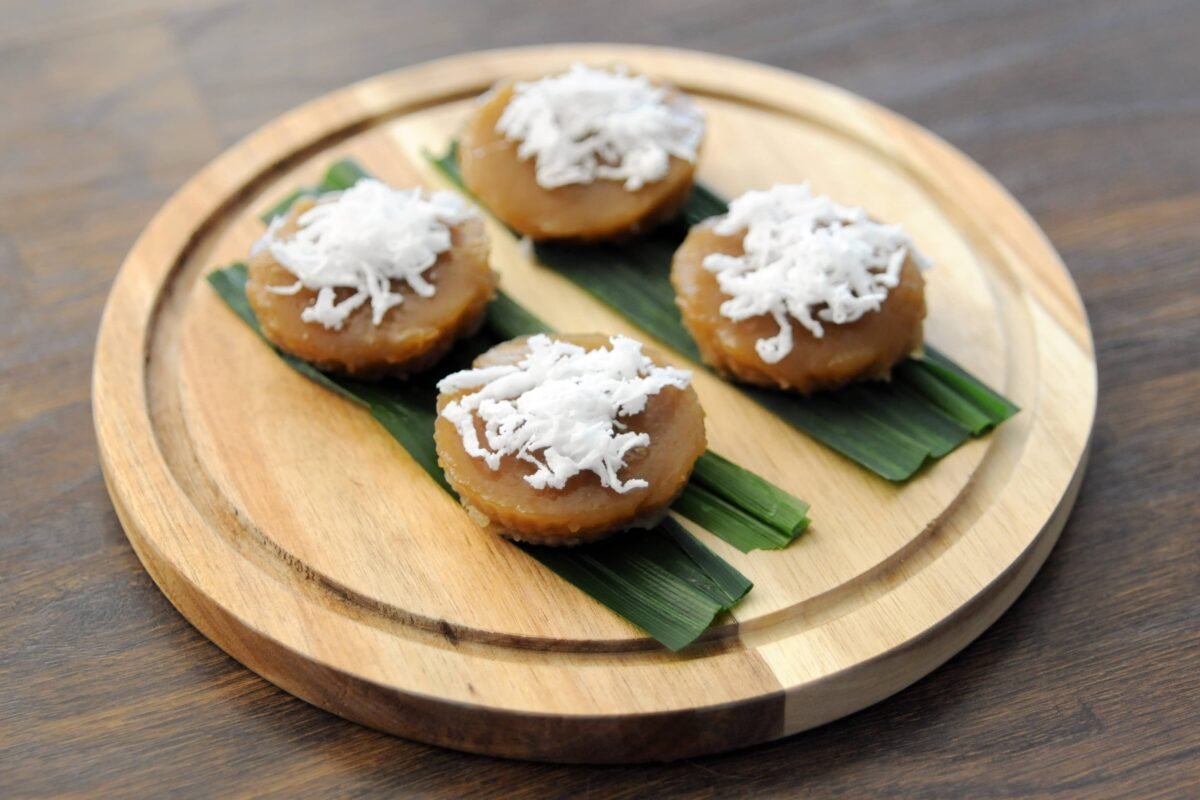 The image displays four pieces of puto kutsinta, a traditional Filipino rice cake, served on top of banana leaves and garnished with shredded coconut. The cakes are presented on a round wooden platter, showcasing their caramel-like color and soft, gelatinous texture.