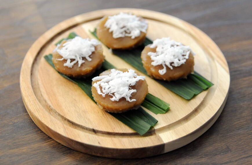 The image displays four pieces of puto kutsinta, a traditional Filipino rice cake, served on top of banana leaves and garnished with shredded coconut. The cakes are presented on a round wooden platter, showcasing their caramel-like color and soft, gelatinous texture.