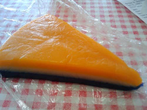 The image shows a slice of Sapin-Sapin, a colorful layered Filipino rice cake. The slice has an orange top layer, a white middle layer, and a dark blue base, all set against a red and white checkered background.