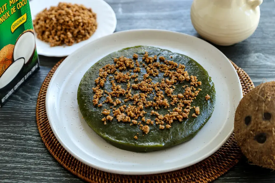 A traditional Filipino Inkiwar Rice Cake topped with a generous sprinkle of sweet coconut crumbs, served on a white plate. The Inkiwar rice cake, with its distinct green hue, is made from glutinous rice and coconut milk, offering a soft and chewy texture, perfect for any festive celebration.