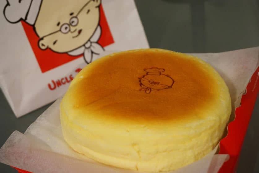 A soft and fluffy Rice Cooker Japanese Cheesecake, perfectly golden on top, with a smooth texture. The cheesecake is presented on parchment paper, featuring a cute chef logo stamped on the surface.