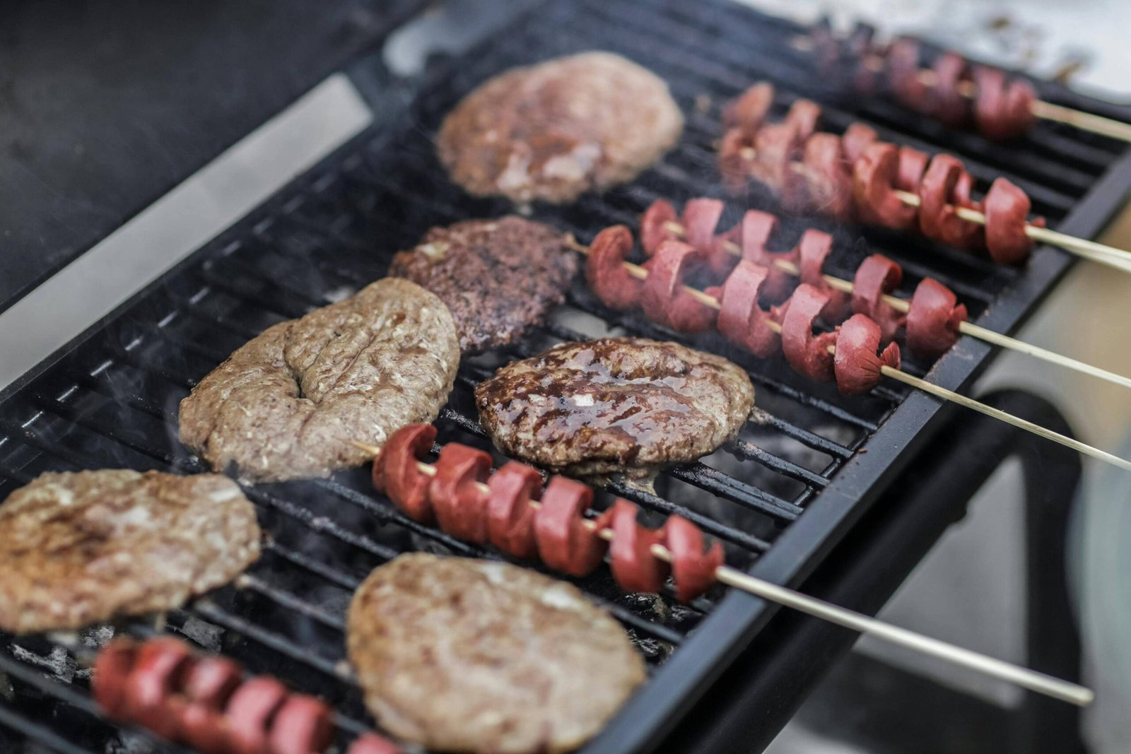 Burger patties and hotdog skewers grilling on a barbecue.