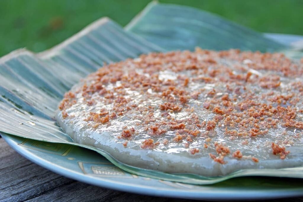 A freshly prepared Kalamay, a sticky and sweet Filipino rice cake, served on a banana leaf. The dessert is topped with a generous sprinkle of toasted coconut bits, adding texture and flavor to the smooth, glossy surface of the rice cake.