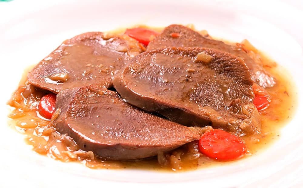 A savory dish of "Lengua Estofado," featuring tender slices of ox tongue cooked in a rich tomato-based sauce with carrots and onions, served on a white plate.