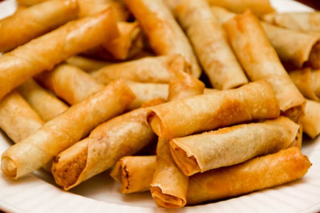 A plate piled with golden-brown, crispy Lumpiang Kamote, which are Filipino spring rolls filled with sweet potato, freshly fried and served on a white plate.