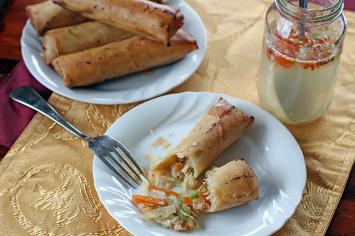 A plate of Lumpiang Sardinas with Sawsawan, featuring crispy spring rolls filled with sardines and vegetables, served alongside a fork and a jar of dipping sauce.