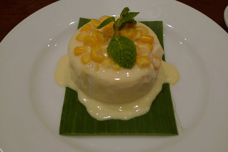 A circular Maja Blanca dessert served on a banana leaf, garnished with sweet corn kernels, mint leaves, and creamy sauce.