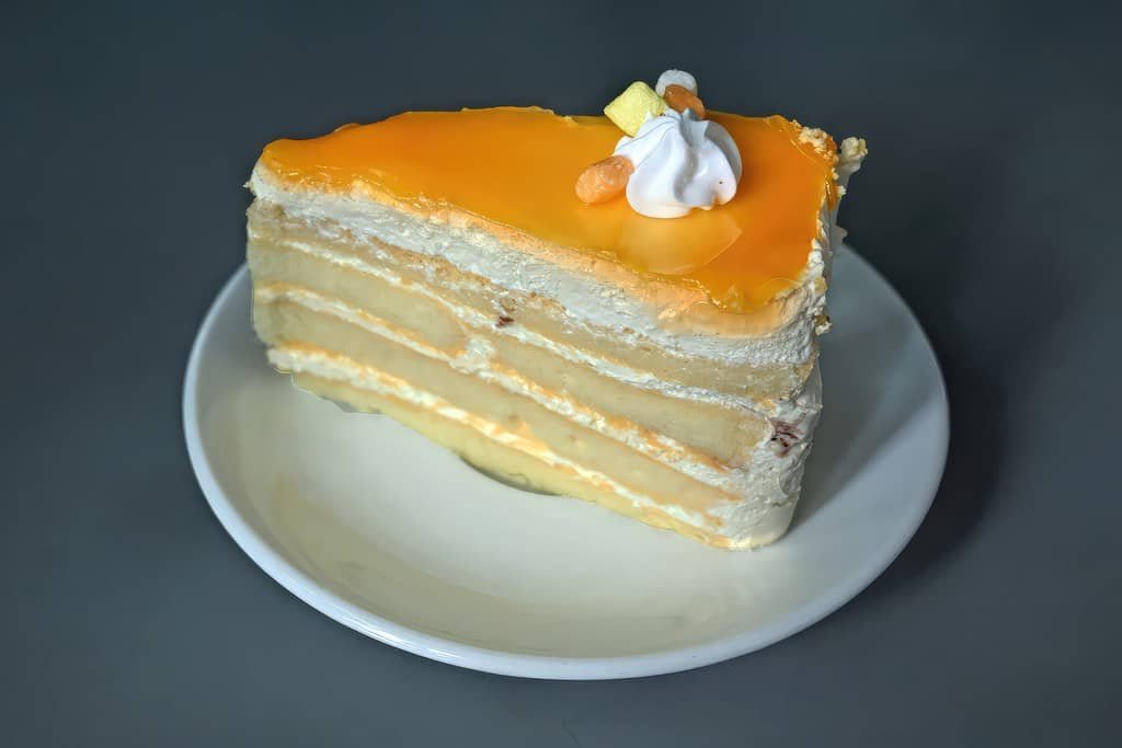 A slice of Mango Cake featuring multiple layers of soft sponge cake filled with cream and topped with a glossy mango-flavored glaze. The cake is garnished with a swirl of whipped cream and small pieces of fresh mango, creating a delightful tropical treat.