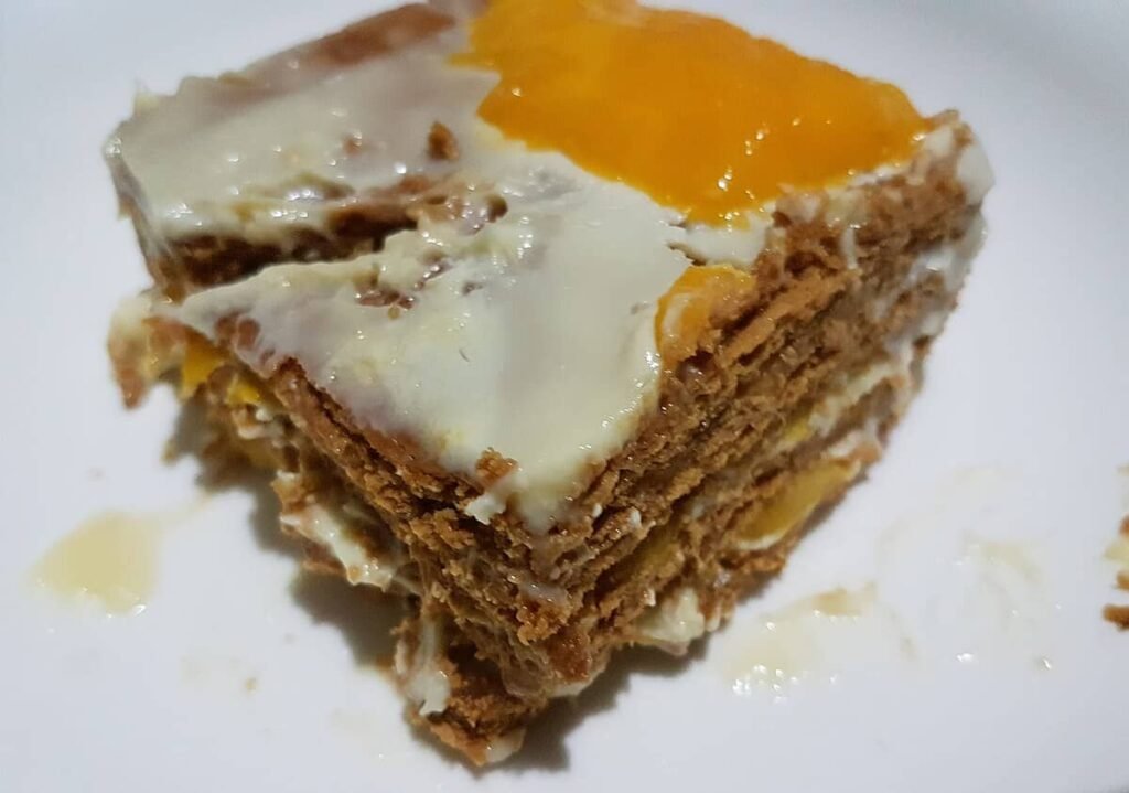 A close-up of a slice of Mango Cream Cheesecake, featuring layers of graham crackers, mango slices, and a creamy cheesecake filling. The top is adorned with a glossy mango topping