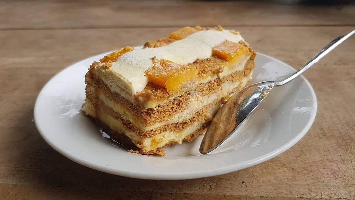 A neatly plated slice of Mango Cream Cheesecake with layers of graham crackers, mango, and cheesecake cream. The dessert is topped with mango slices and served with a spoon, resting on a wooden table.