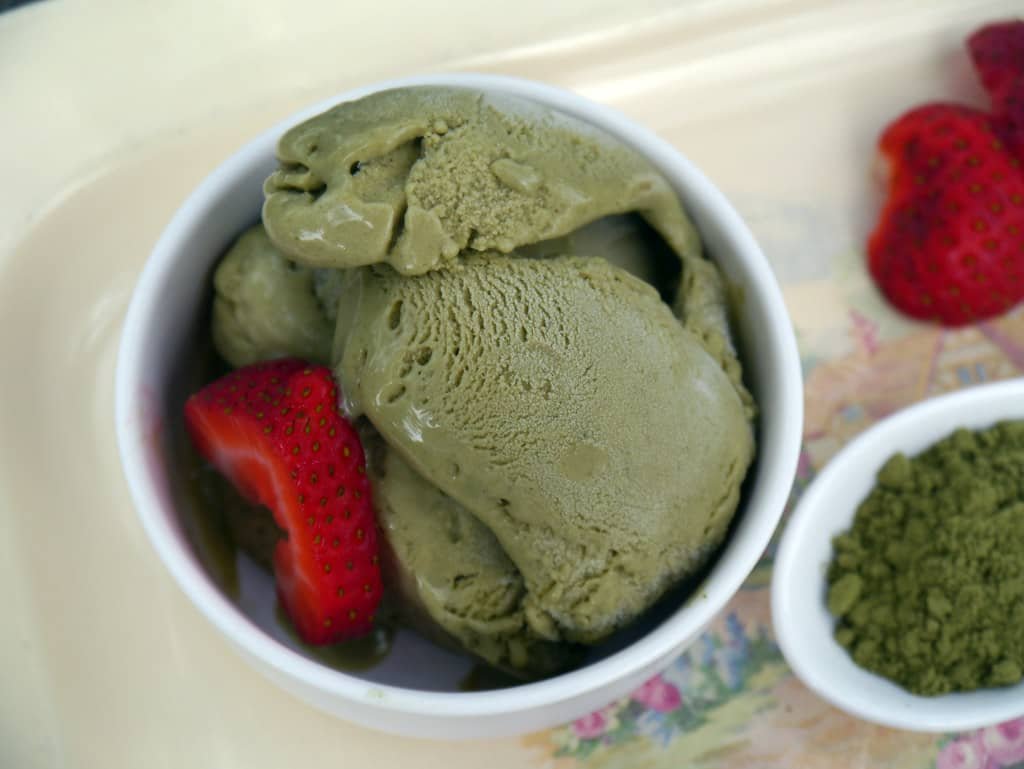 A bowl of creamy matcha ice cream served with fresh strawberry slices, alongside a small dish of matcha powder.