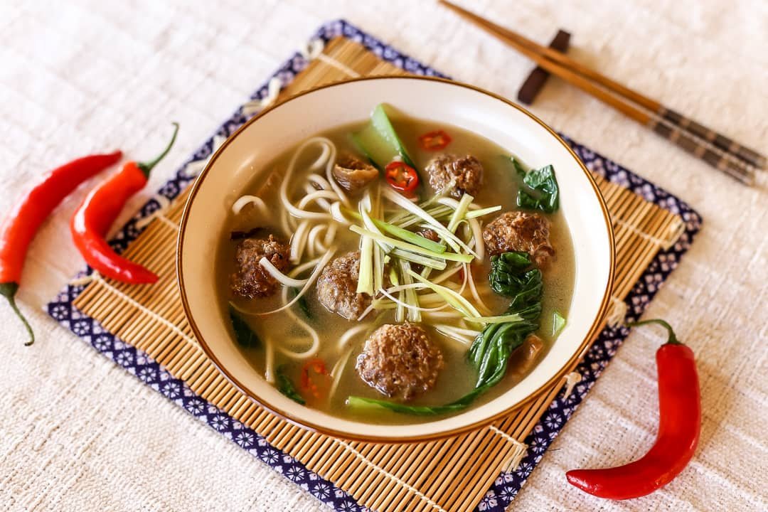 A bowl of Miswa and Meat Balls Soup Recipe with noodles, meatballs, fresh vegetables, and a light broth garnished with chili peppers, served on a traditional mat.