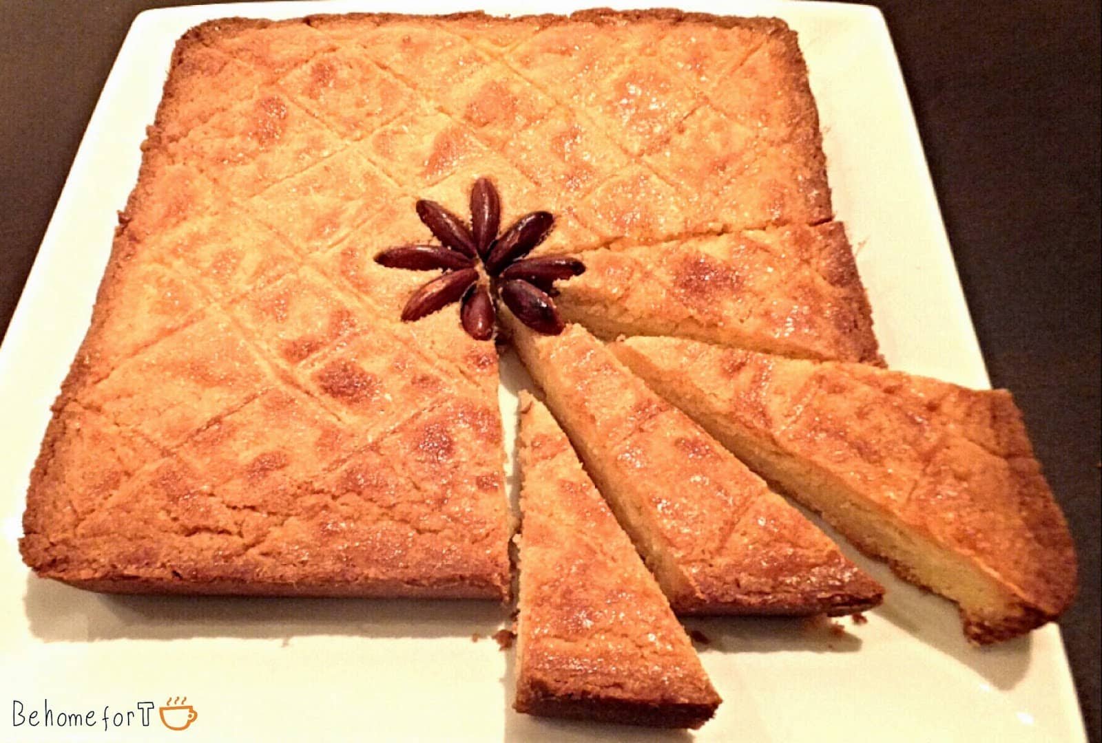 "A golden-brown, moist butter cake displayed on a white plate, cut into triangular slices. The cake has a crisscross pattern on the surface, and an anise star is placed in the center for decoration, enhancing the presentation.