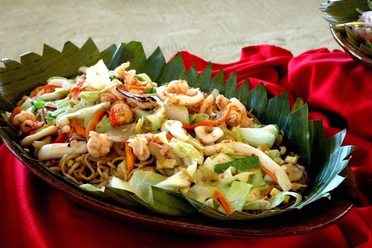 The image features a delicious and vibrant platter that highlights seafood pancit, a popular Filipino dish that is a must-try in Davao. It showcases fresh seafood like shrimp, squid, and vegetables such as cabbage and carrots, mixed with pancit noodles and seasoned with spices. The dish is served on banana leaves, adding a rustic and authentic touch typical of Filipino cuisine.