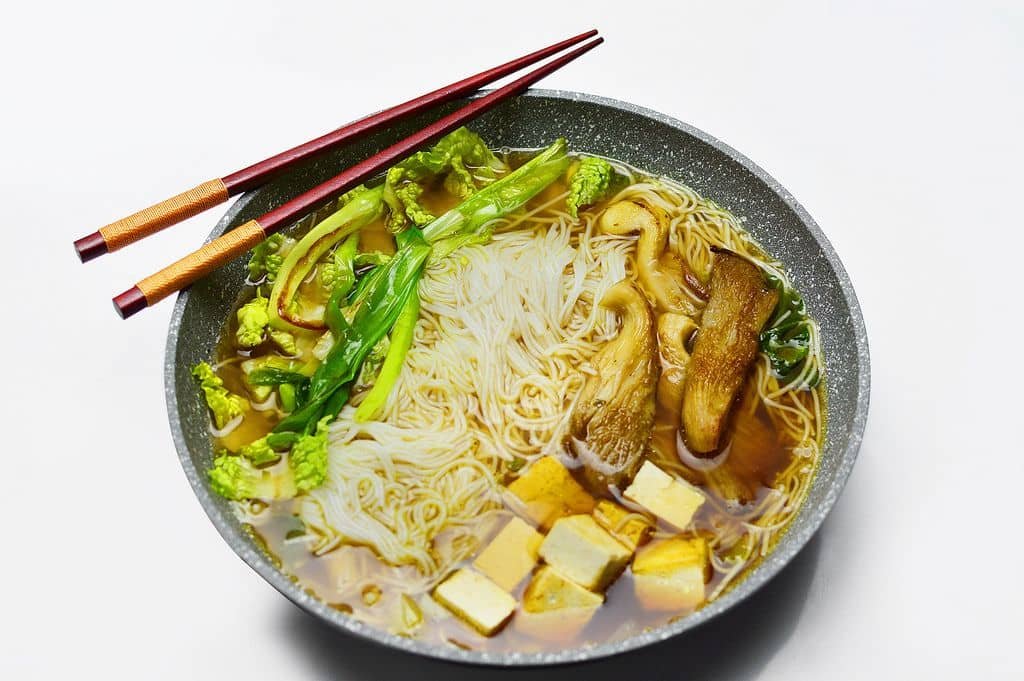 A bowl of Norwood Chinese Restaurant Soup, featuring a rich broth with delicate rice noodles, large slices of king oyster mushrooms, and fresh greens. Tofu cubes and leafy vegetables float in the aromatic soup, with a pair of red chopsticks resting elegantly on the bowl’s edge, highlighting the dish's vibrant and inviting presentation.