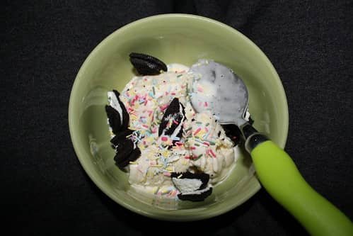 A bowl of Oreo Ice Candy topped with crushed Oreo cookies and colorful sprinkles, featuring a smooth and creamy frozen dessert made with milk and Oreo chunks, served in a simple green dish with a green-handled spoon.